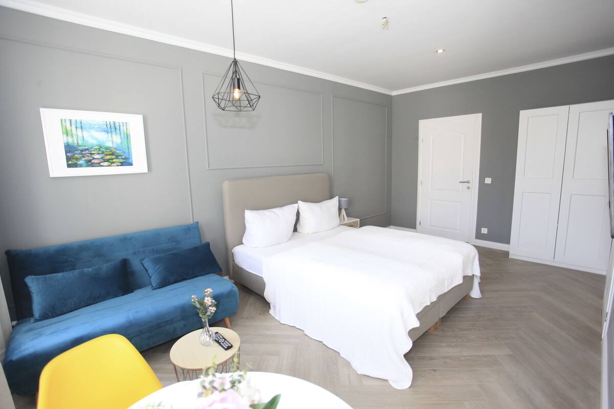 Un dormitorio con una cama, un sofá azul, un sillón amarillo, una mesita redonda con un florero y un cuadro en la pared.