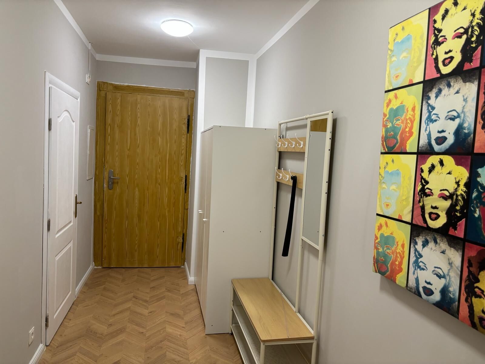 A hallway with a wooden door, a white refrigerator, a bench, and a pop art mural on the wall.