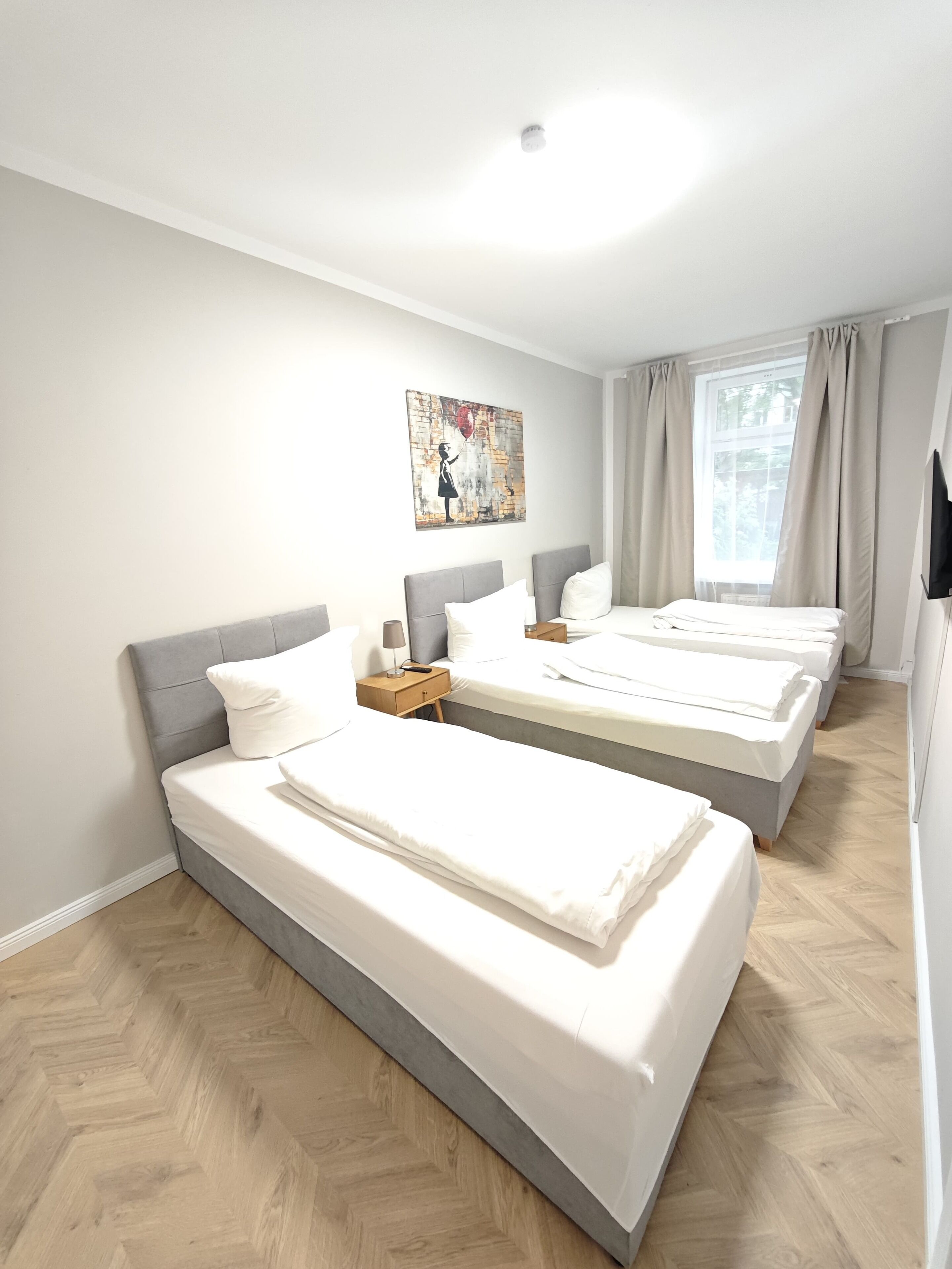 A hotel room with three single beds, a wooden herringbone floor, a painting on the wall, and a window with curtains.