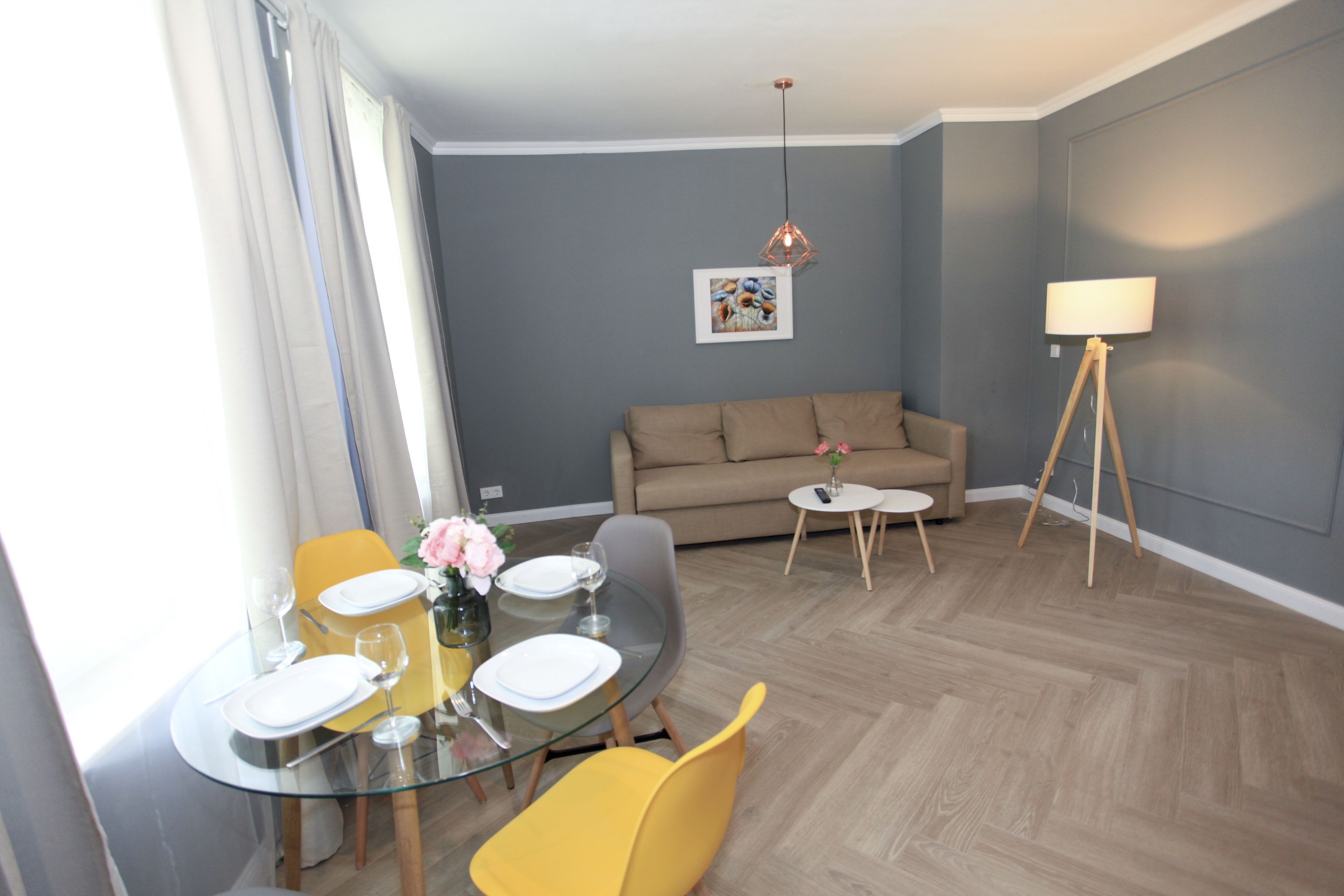 Una sala de estar moderna con una mesa de comedor redonda de vidrio, sillas amarillas, un sofá color beige, una lámpara de pie y un cuadro en la pared.