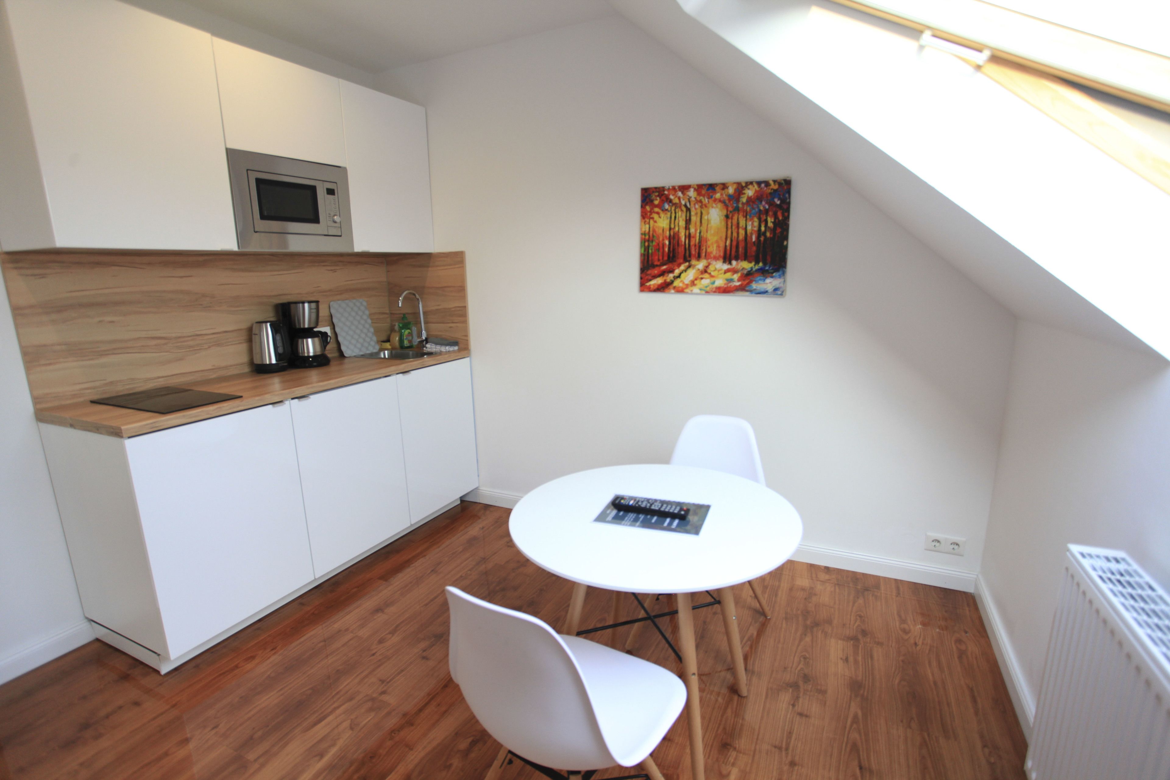 Eine moderne Küche mit weißen Schränken, einer Mikrowelle und einer Kaffeemaschine. Ein weißer Tisch steht dort mit einem Stuhl, und an der Wand hängt ein Bild.