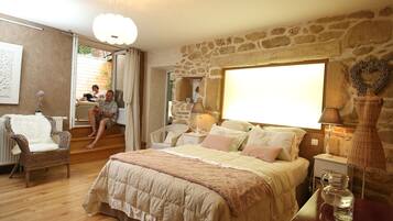 A bedroom with a stone wall, a large bed with multiple pillows, a wicker chair, and a bedside table with a lamp.