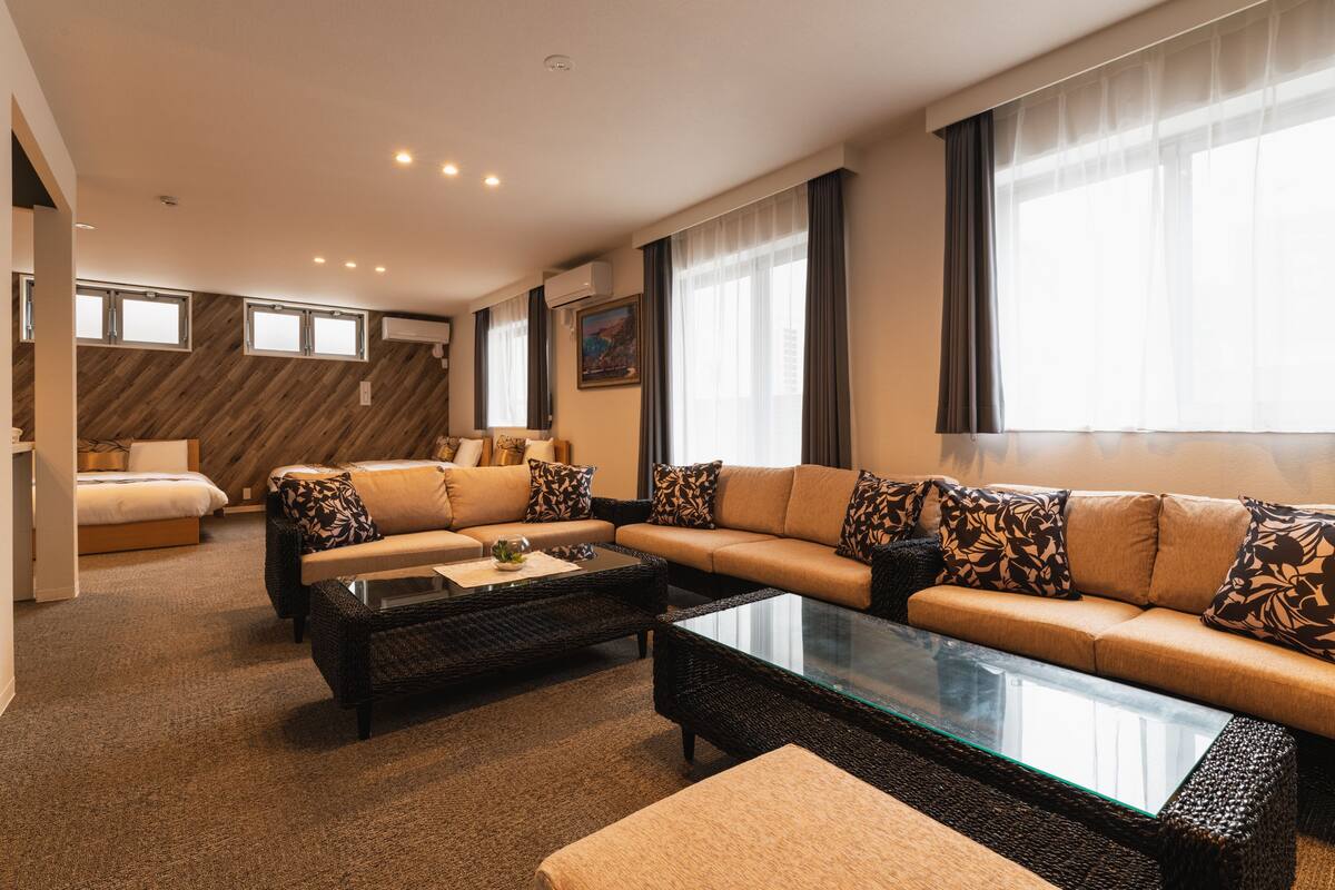 A modern hotel room with a large sofa set, a glass coffee table, and a bed with a wooden headboard.