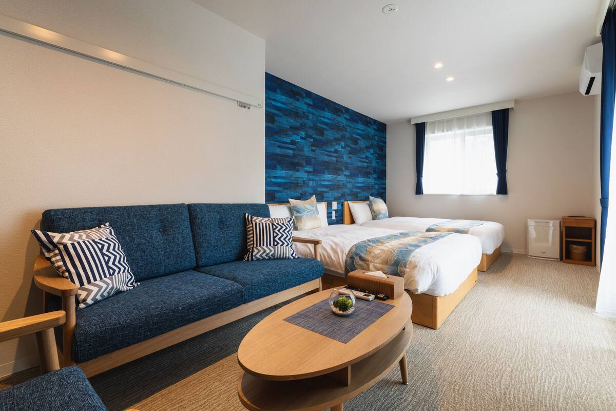 A modern hotel room with a blue sofa, a large bed, a wooden coffee table, and a blue accent wall.