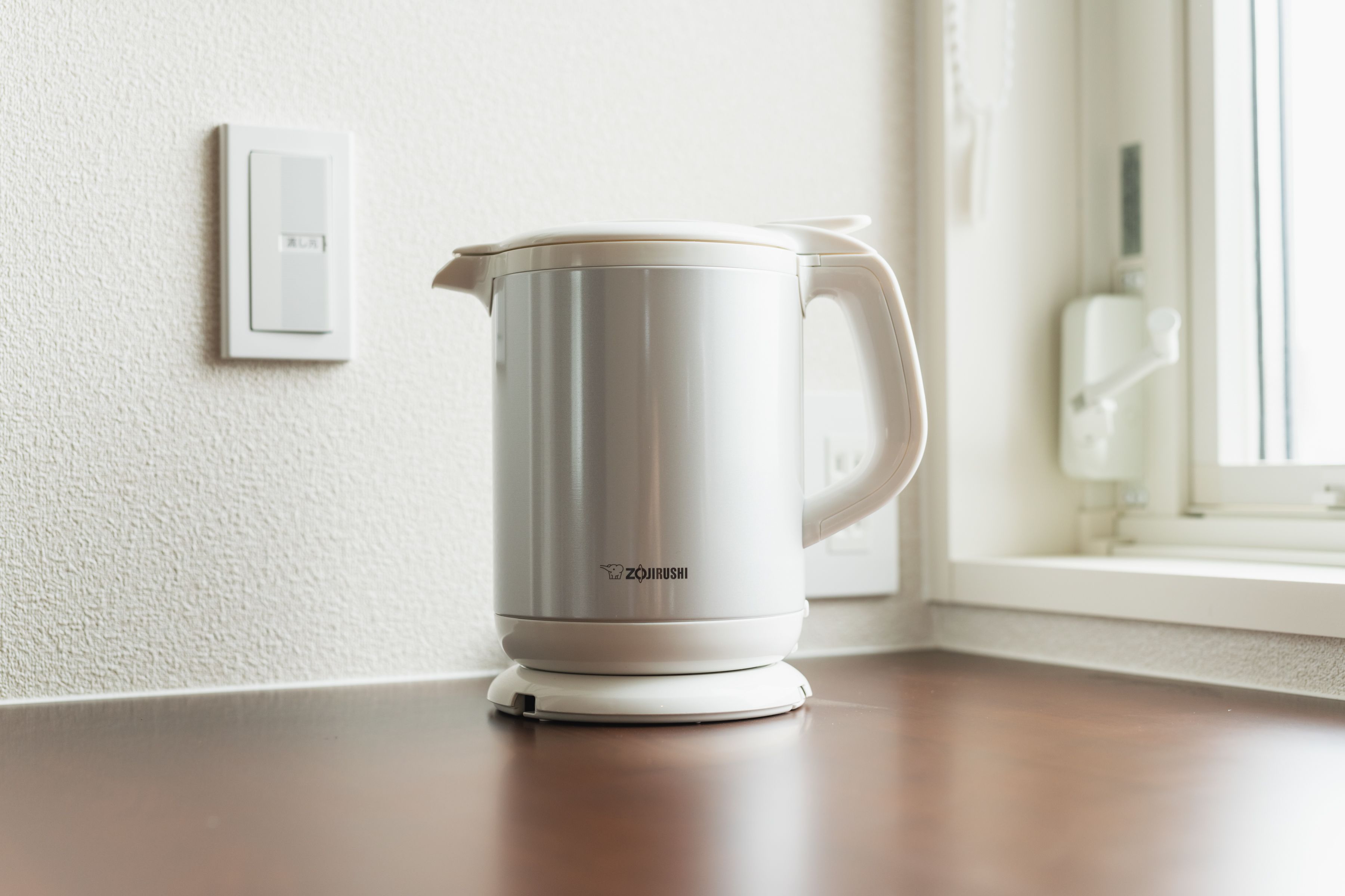 A white electric kettle on a wooden countertop near a window.