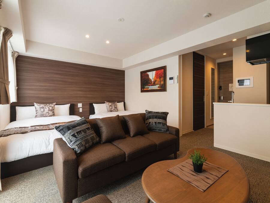 Una habitación de hotel moderna con una cama, un sofá marrón, una mesa de centro redonda y un televisor montado en la pared.