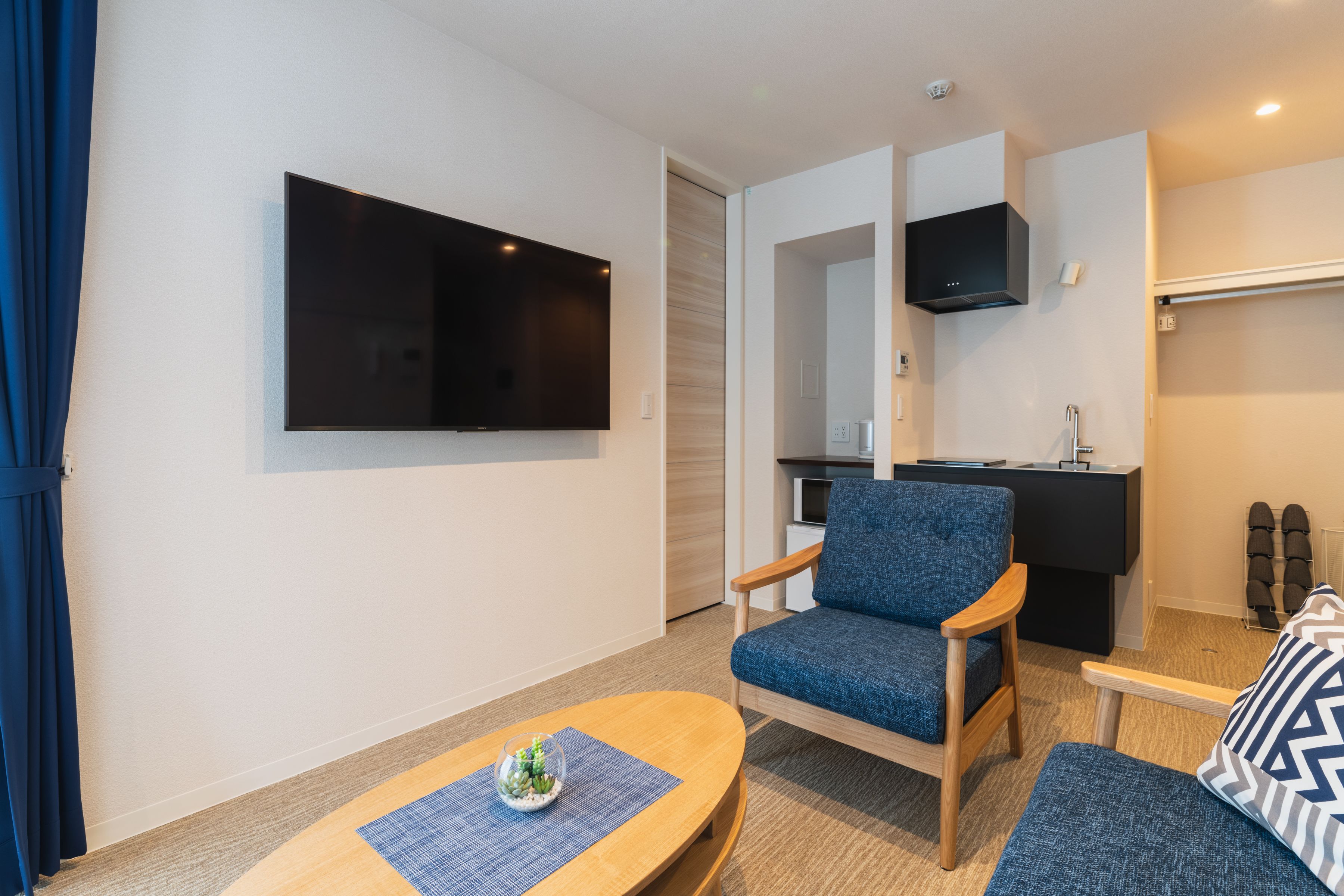 A modern living room with a flat-screen TV, a blue armchair, a wooden coffee table, and a small kitchen area with a black countertop and a microwave.