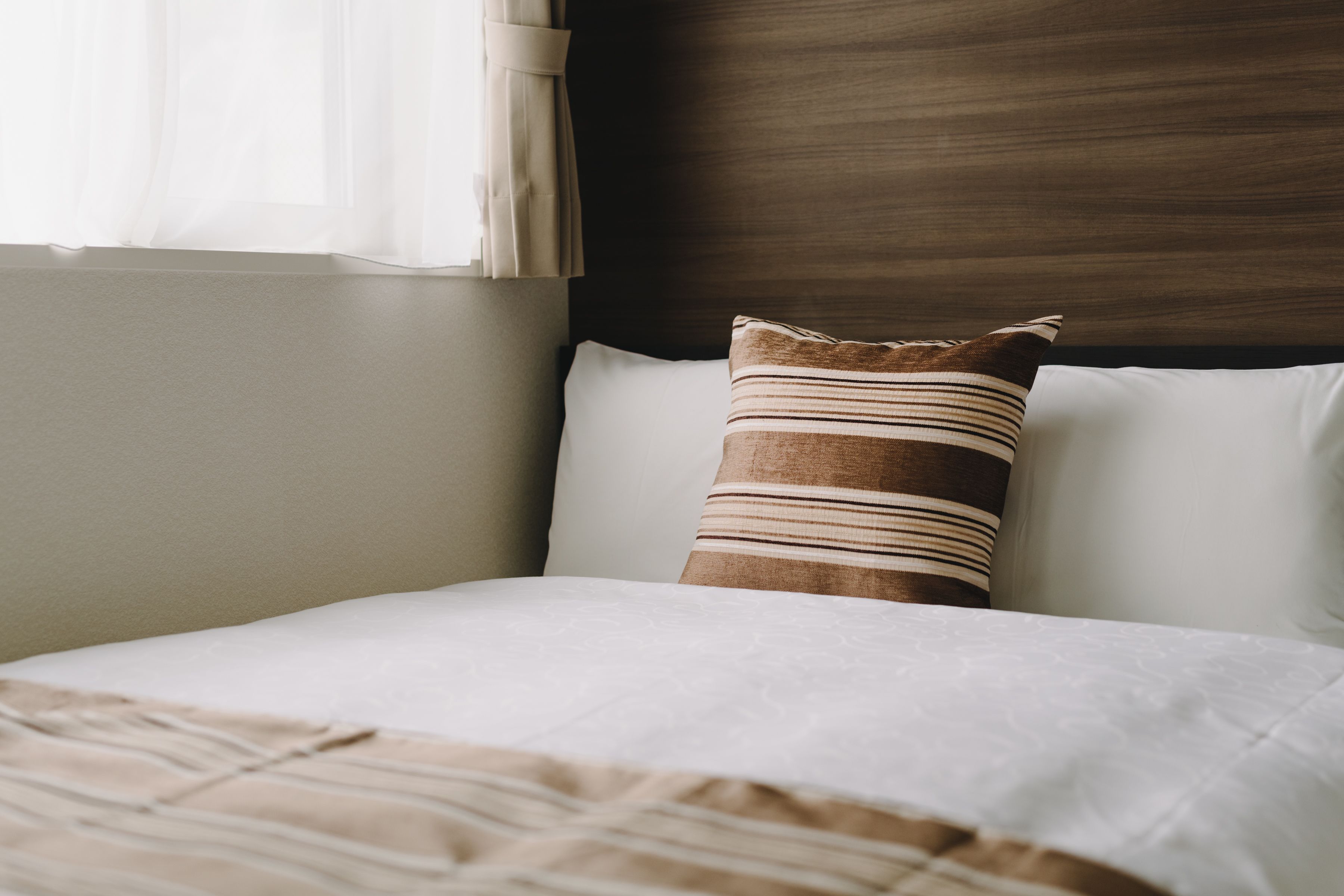 Cama bem arrumada com travesseiro listrado e travesseiro branco liso, cabeceira de madeira e janela com cortinas translúcidas.