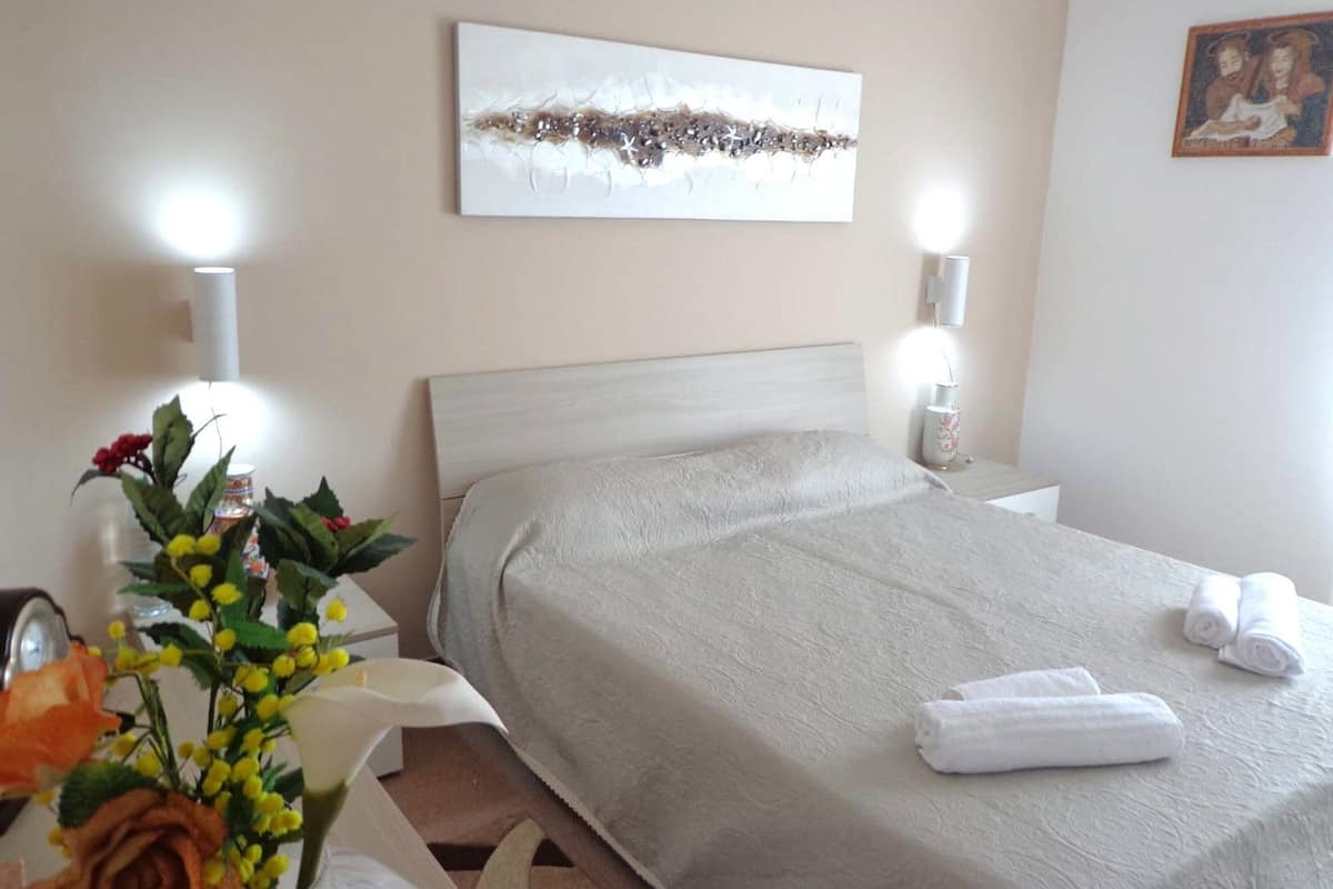 A hotel room with a bed, bedside tables, a lamp, a vase with flowers, and a painting on the wall.