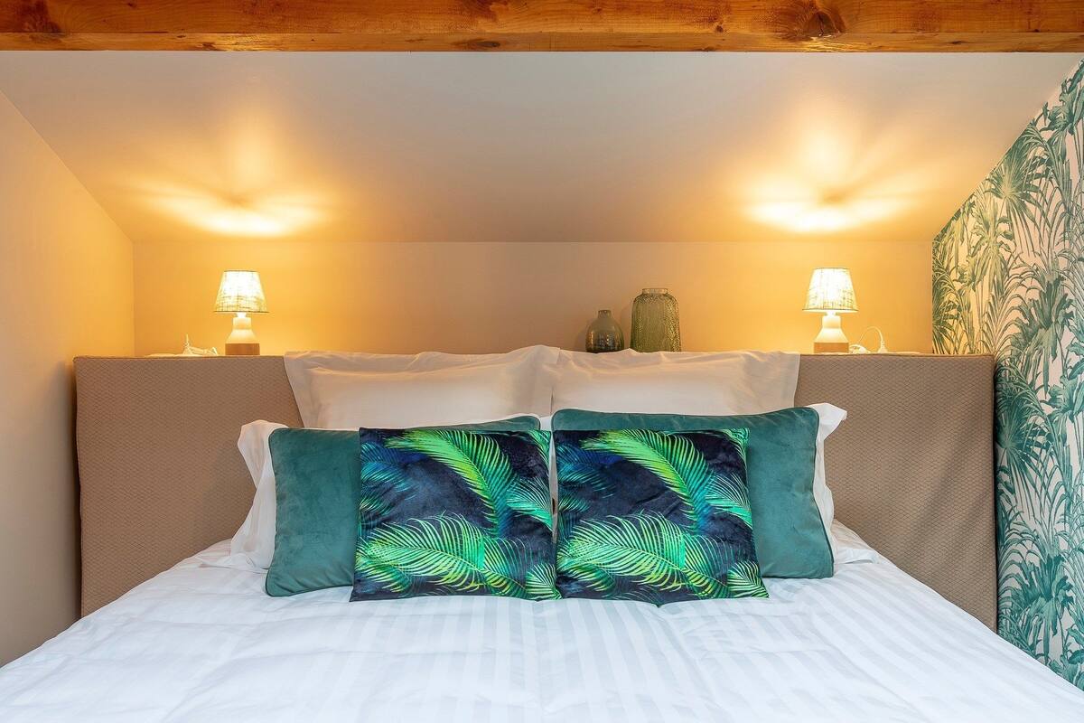 A bed with white linens, two green pillows with a tropical leaf pattern, and two bedside lamps on a wooden headboard.