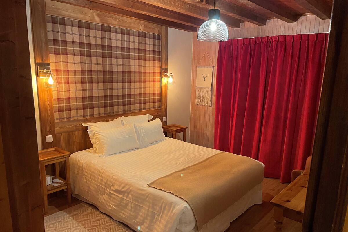 A cozy bedroom with a large bed, wooden headboard, plaid curtains, and a hanging light fixture.
