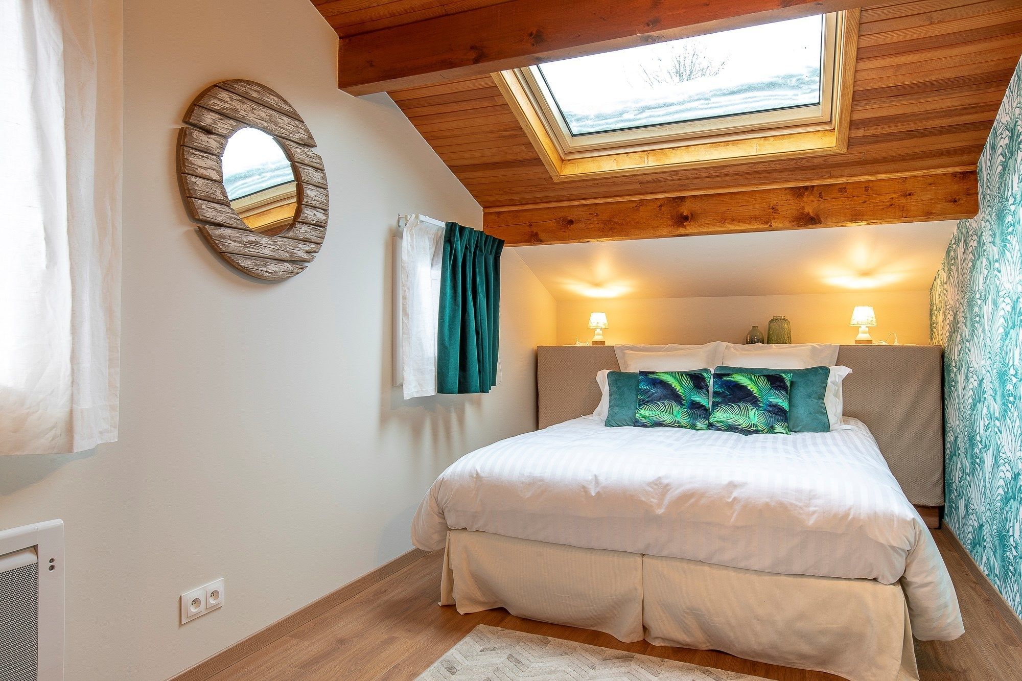 A bedroom with a skylight, a round mirror, a bed with white bedding, and a patterned wall.