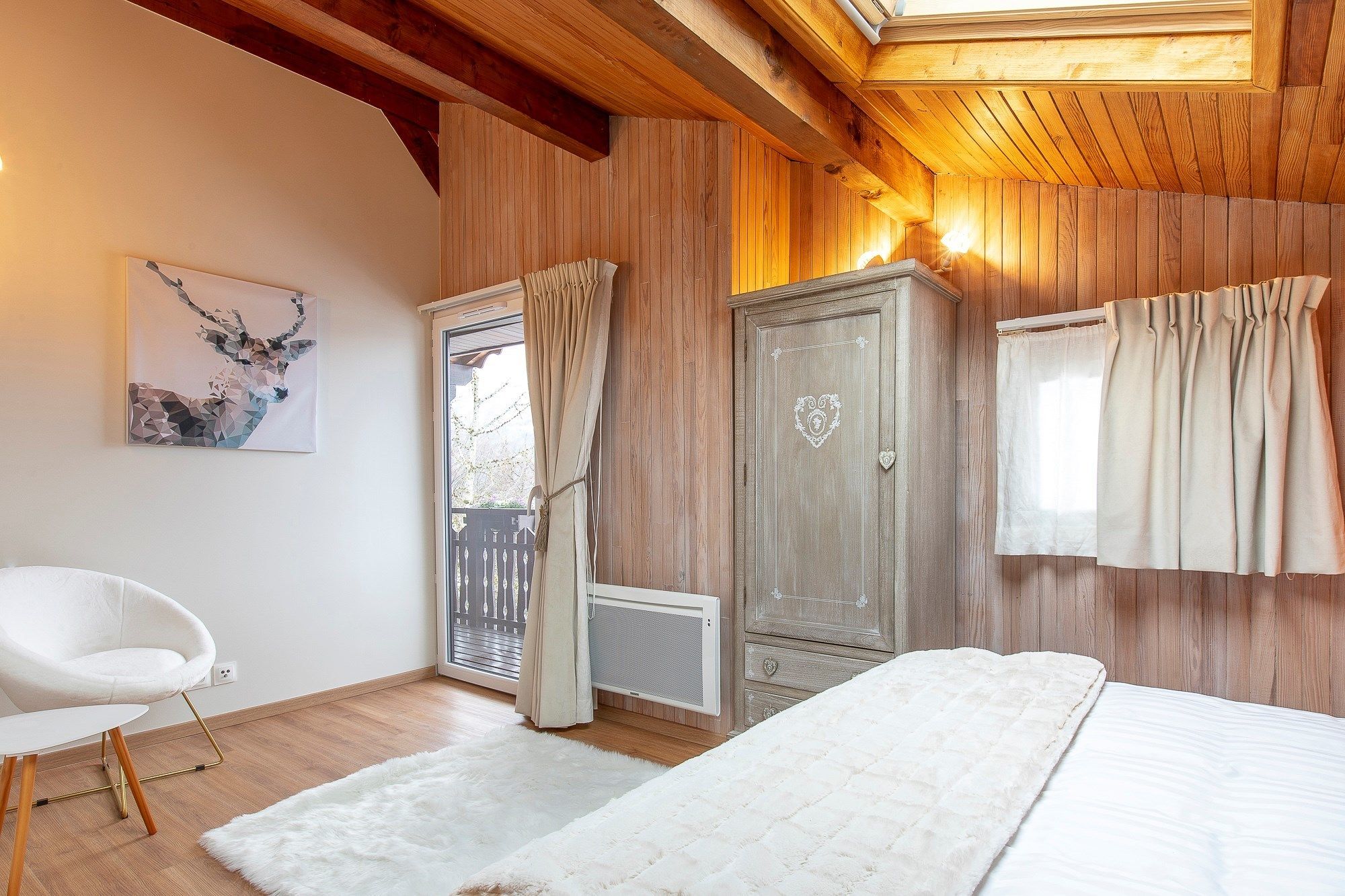 Une chambre avec un plafond en bois, une chaise blanche, un lit recouvert d’une couverture blanche, une armoire et une fenêtre avec des rideaux.