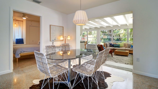 A modern dining area with a glass table and wicker chairs, a ceiling fan, and a view of a living room with a sofa and large windows.