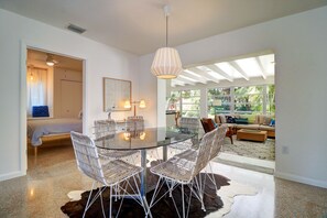 Un espace repas moderne avec une table en verre et des chaises en osier, un ventilateur de plafond et une vue sur un salon doté d’un canapé et de grandes fenêtres.