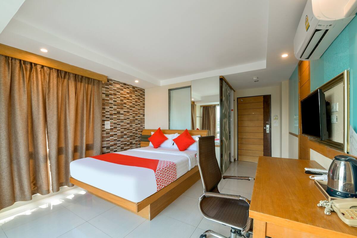 A hotel room with a bed, a desk with a kettle, a chair, and a TV.