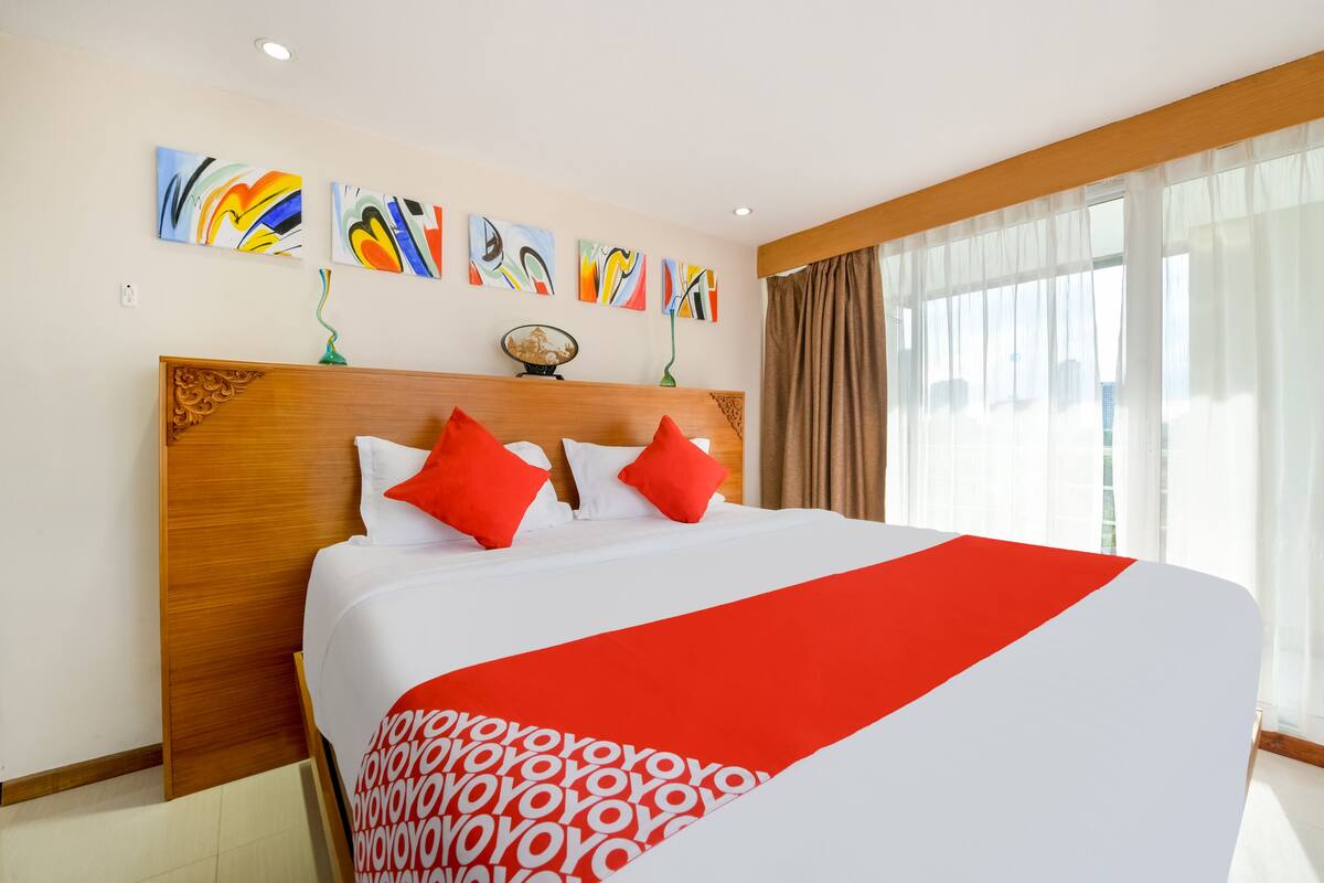 A hotel room with a large bed, red and white bedding, a wooden headboard, and abstract art on the wall.