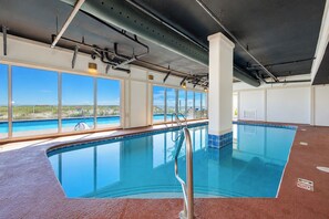 Une piscine intérieure avec de grandes baies vitrées, offrant une vue sur l’extérieur, et un sol carrelé.