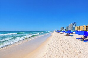 Une plage bordée d’une rangée de parasols bleus et de chaises longues, un rivage sablonneux et un ciel bleu dégagé.