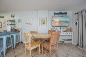 Un coin repas comprenant une table ronde en bois, quatre chaises, une console blanche agrémentée d’objets décoratifs et un tableau représentant une scène de plage.