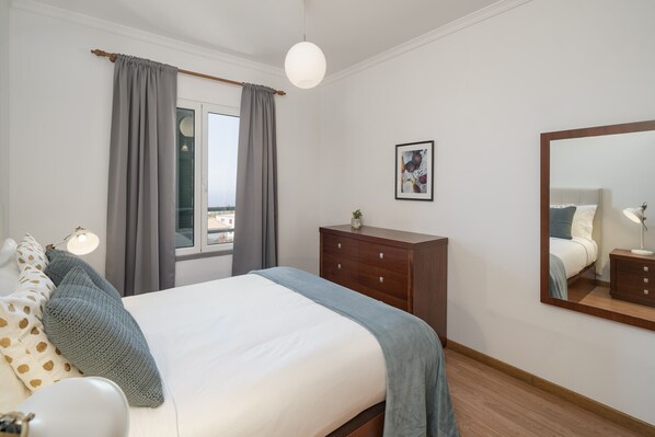A bedroom with a bed, a dresser, a bedside lamp, a window with curtains, a wall-mounted mirror, and a framed picture.