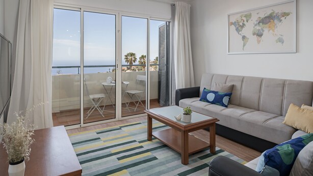 A modern living room with a sofa, a coffee table, and a view of the sea through large glass doors.