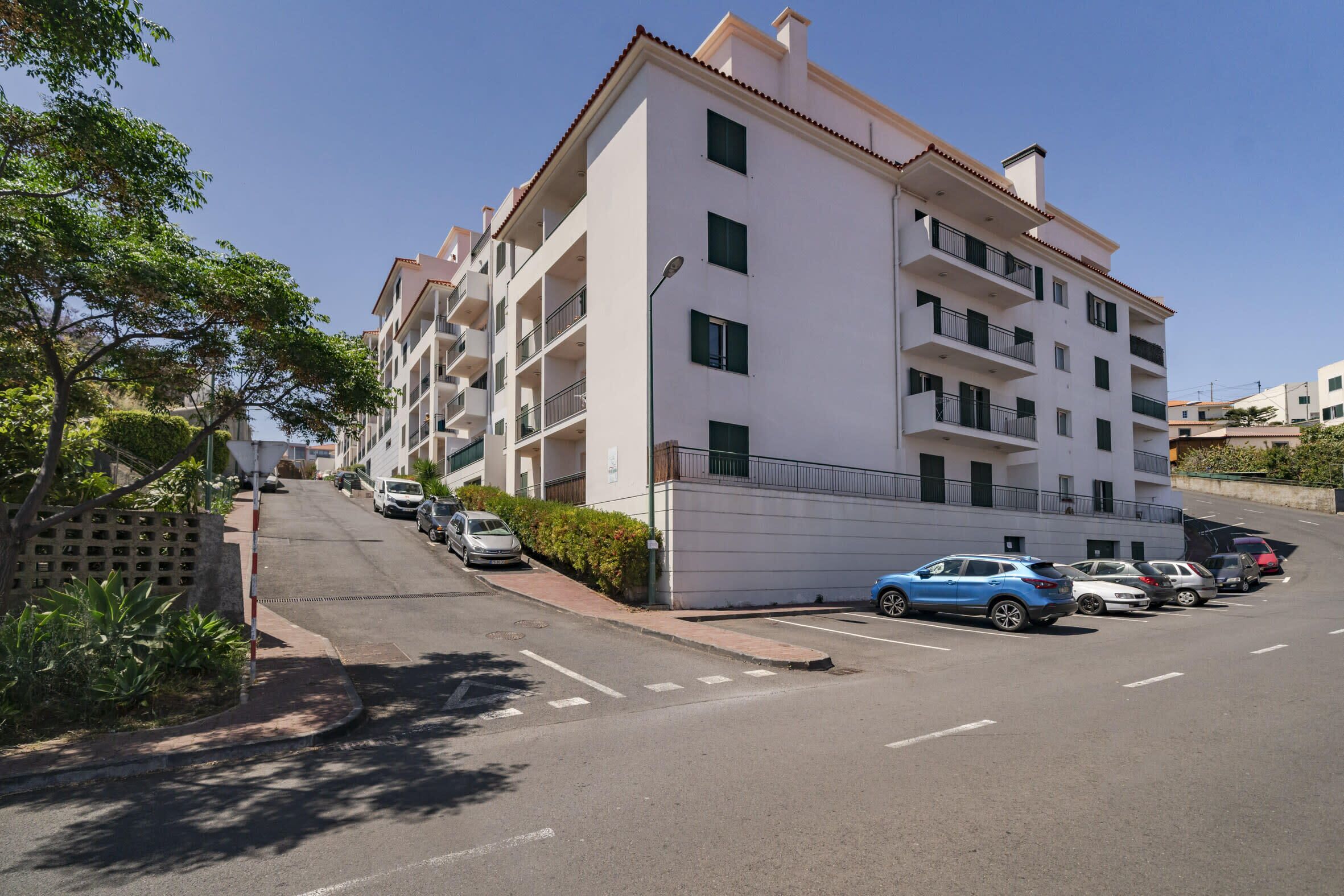Un immeuble résidentiel à plusieurs étages, doté de balcons, avec des voitures garées devant et un ciel dégagé.