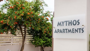 En skylt med texten ”MYTHOS APARTMENTS”, med ett träd och ett staket i bakgrunden.