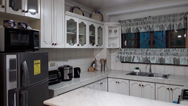 Una cocina con armarios blancos, microondas, refrigerador, cafetera y ventana con cortinas.