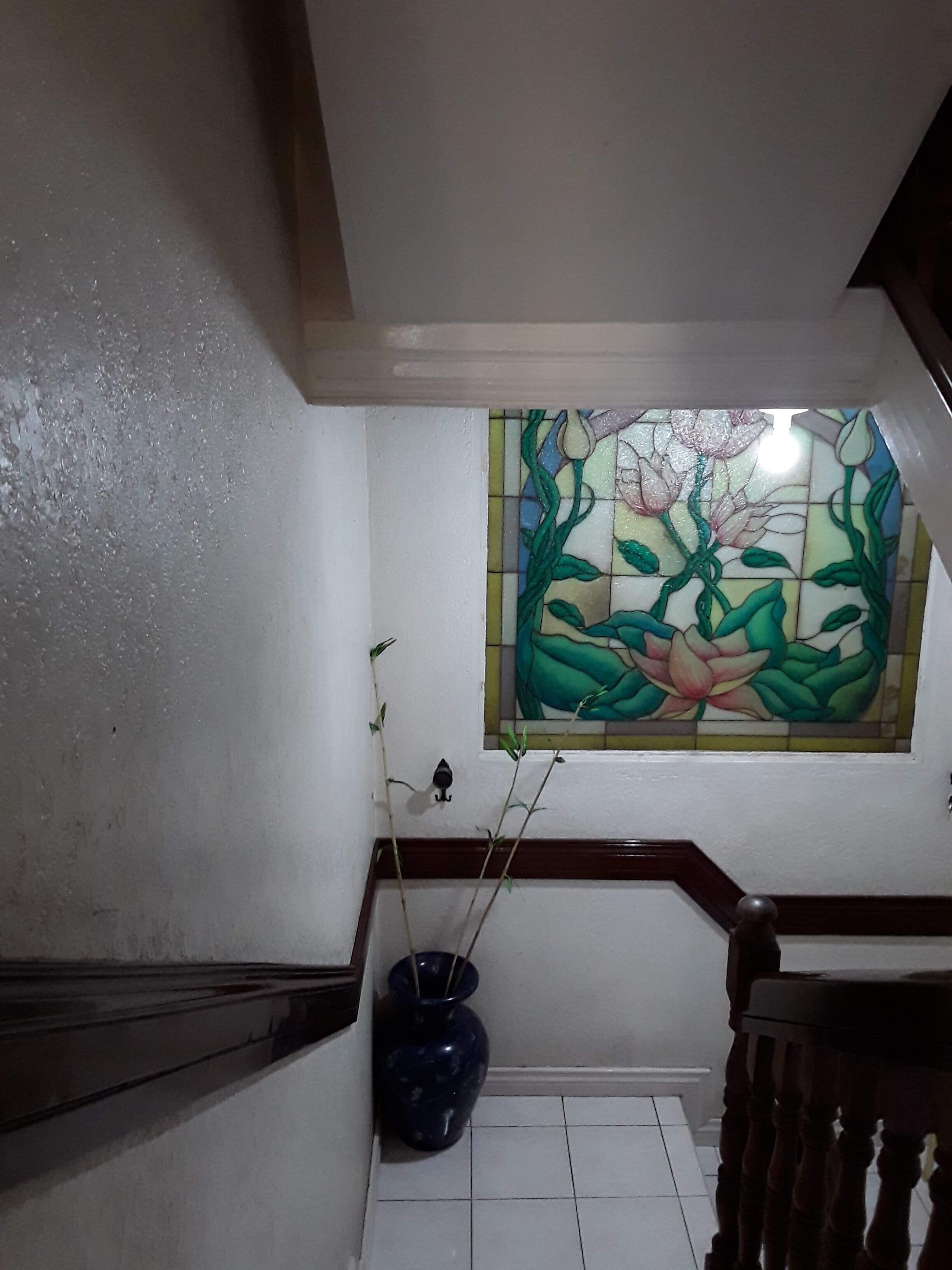 Un escalier avec une rampe en bois, un vase bleu contenant des plantes et une fenêtre à vitraux ornée de motifs floraux.