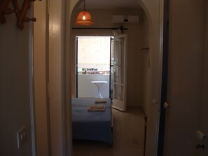 Ein schmaler Flur, der zu einem Schlafzimmer mit einem Bett, einem kleinen Tisch und einer Pendelleuchte führt.