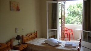 A room with two beds, a nightstand, a lamp, a painting, and a view of greenery through an open door. - Anya Studios (Corfu)