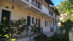 A multi-story building with balconies, a white facade, and a garden area with plants. - Anya Studios (Corfu)