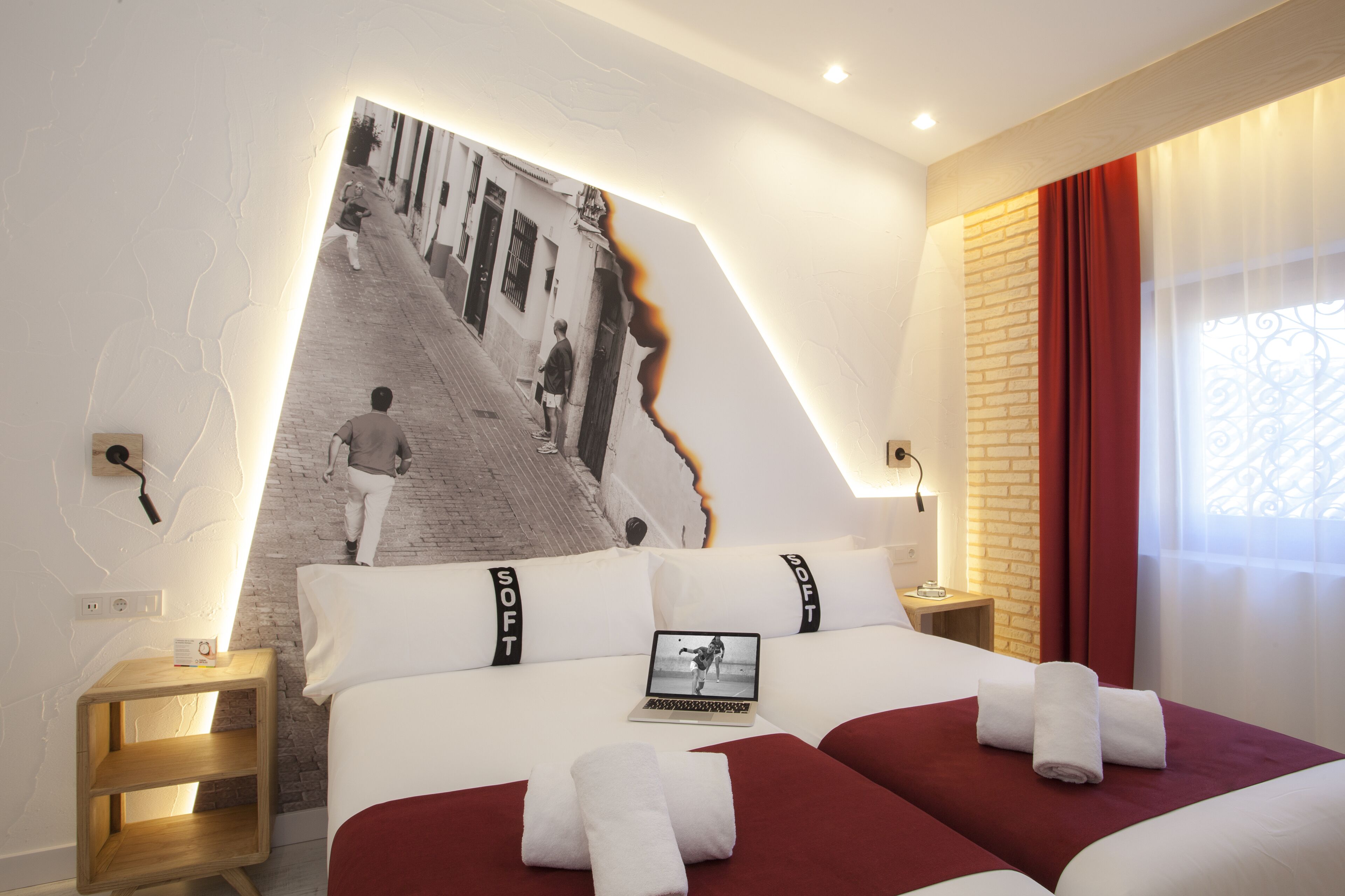 A hotel room with a large wall mural, two beds with red bedding, a laptop on one bed, and a nightstand with a lamp.