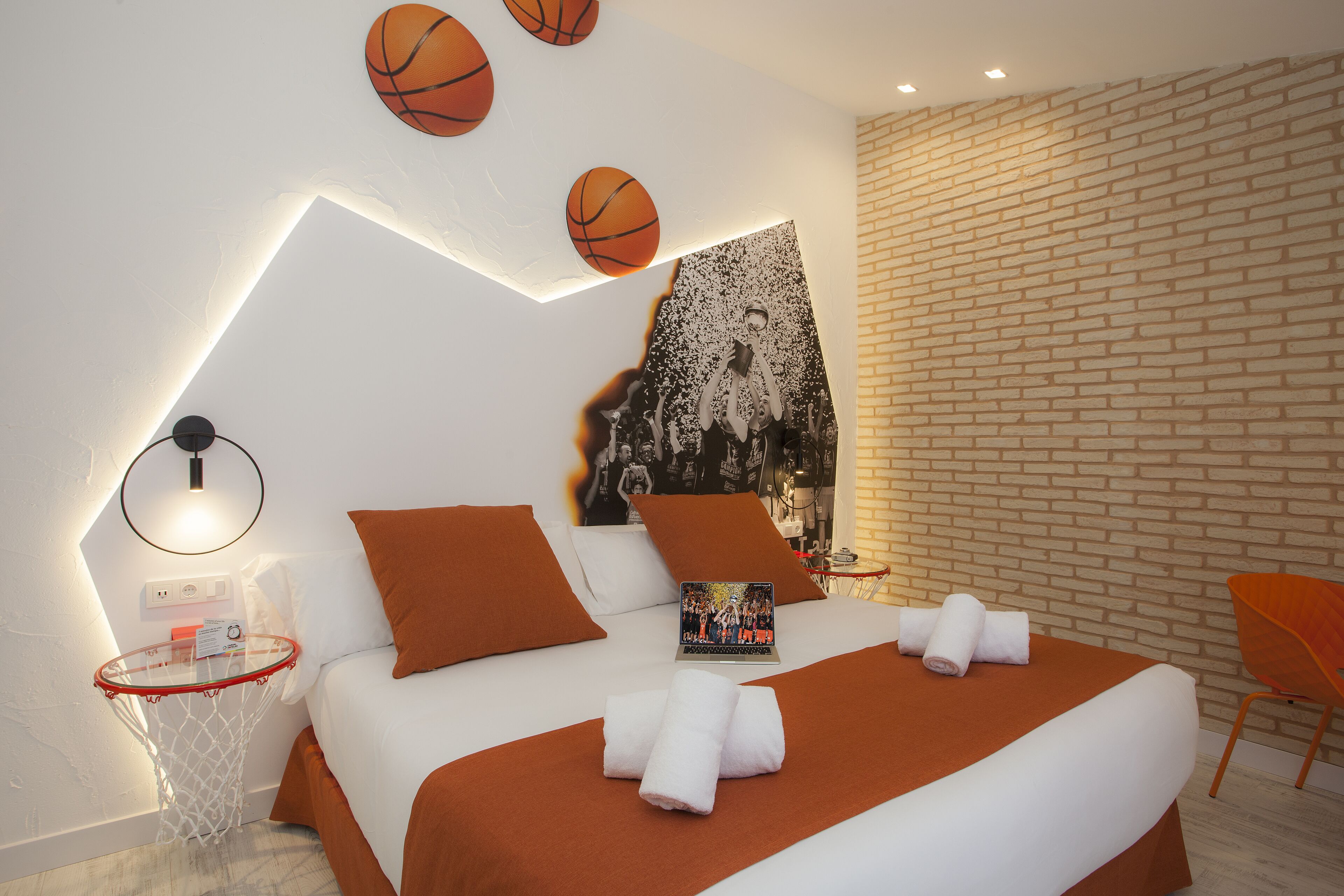 A modern hotel room with a bed, a small round table, a chair, and a basketball-themed wall decoration.