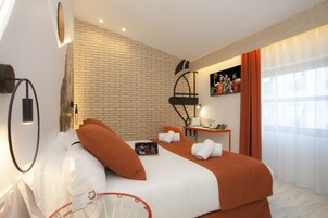 Habitación de hotel con pared de ladrillo, cama con ropa de cama blanca y naranja, mesita de noche con lámpara y ventana con cortinas.