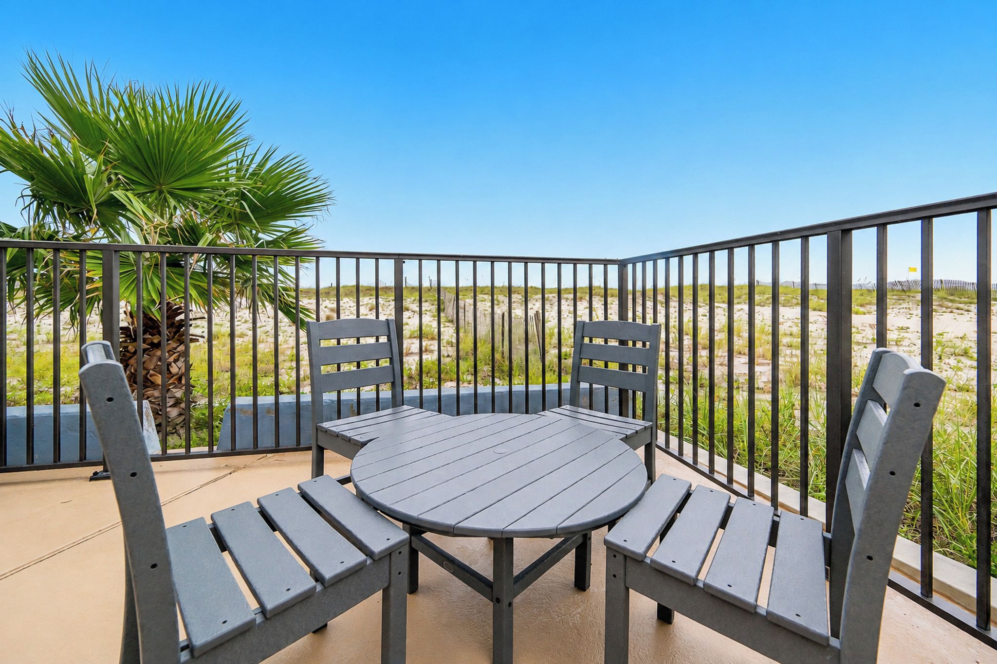 A balcony with a round table and four chairs, surrounded by a metal railing, overlooking a beach with sand dunes and sparse vegetation.