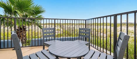 A balcony with a round table and four chairs, surrounded by a metal railing, overlooking a beach with sand dunes and sparse vegetation.