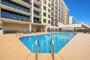 Uma área de piscina de hotel com piscina azul cristalina, espreguiçadeiras e um edifício com várias andares e varandas.
