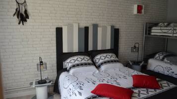 Un dormitorio con pared de ladrillo blanco, cama con ropa de cama estampada y almohadas rojas, mesita de noche con lámpara y un atrapasueños en la pared.