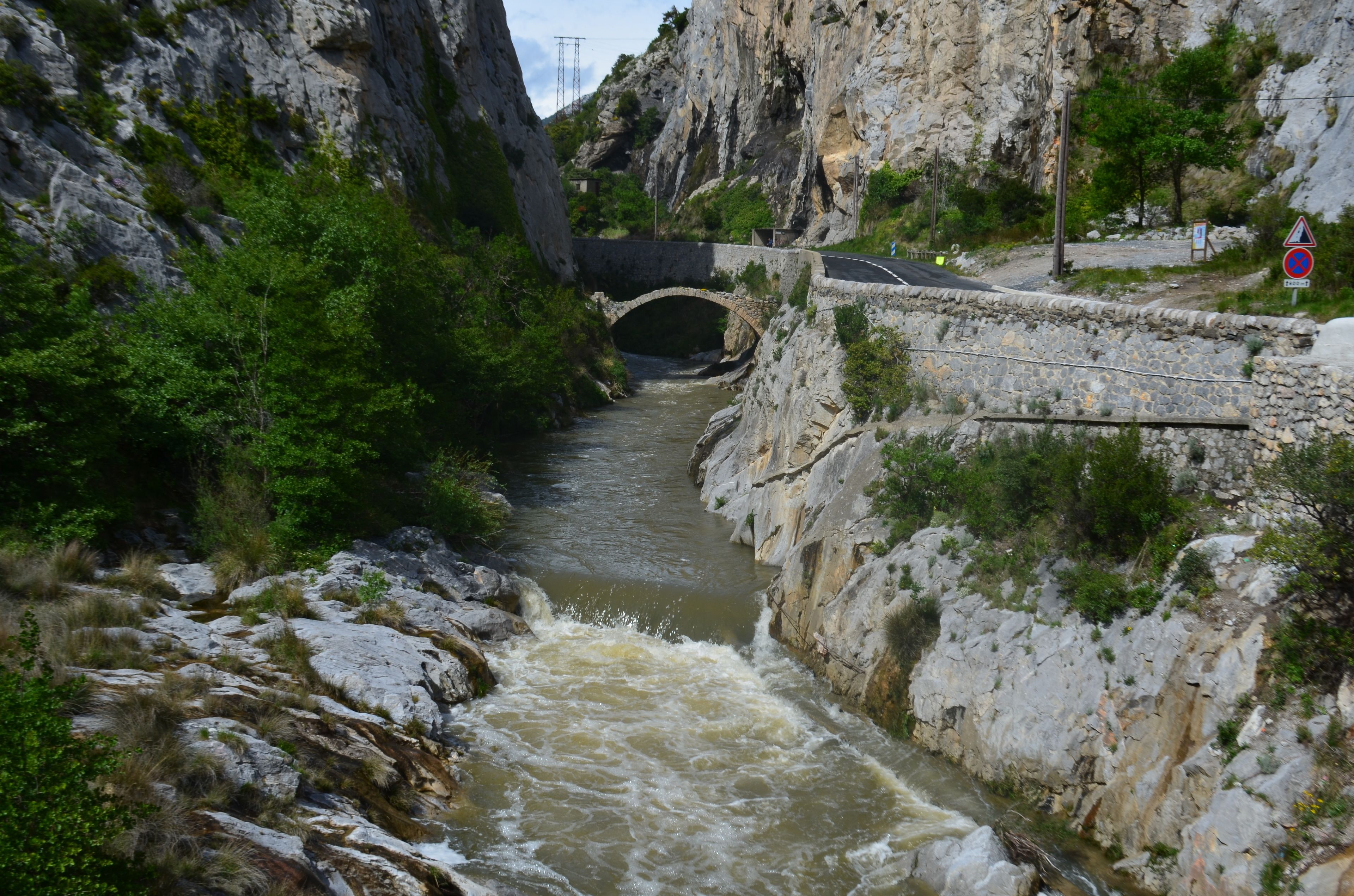 Ein Fluss fließt unter einer Steinbrücke, rechts davon verläuft eine Straße.