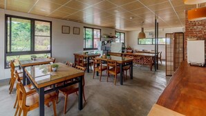 A spacious dining area with multiple wooden tables and chairs, large windows offering views of greenery, and a kitchen area with modern appliances. - Hou Shan Ren Jia B&B Hall A (Luye)