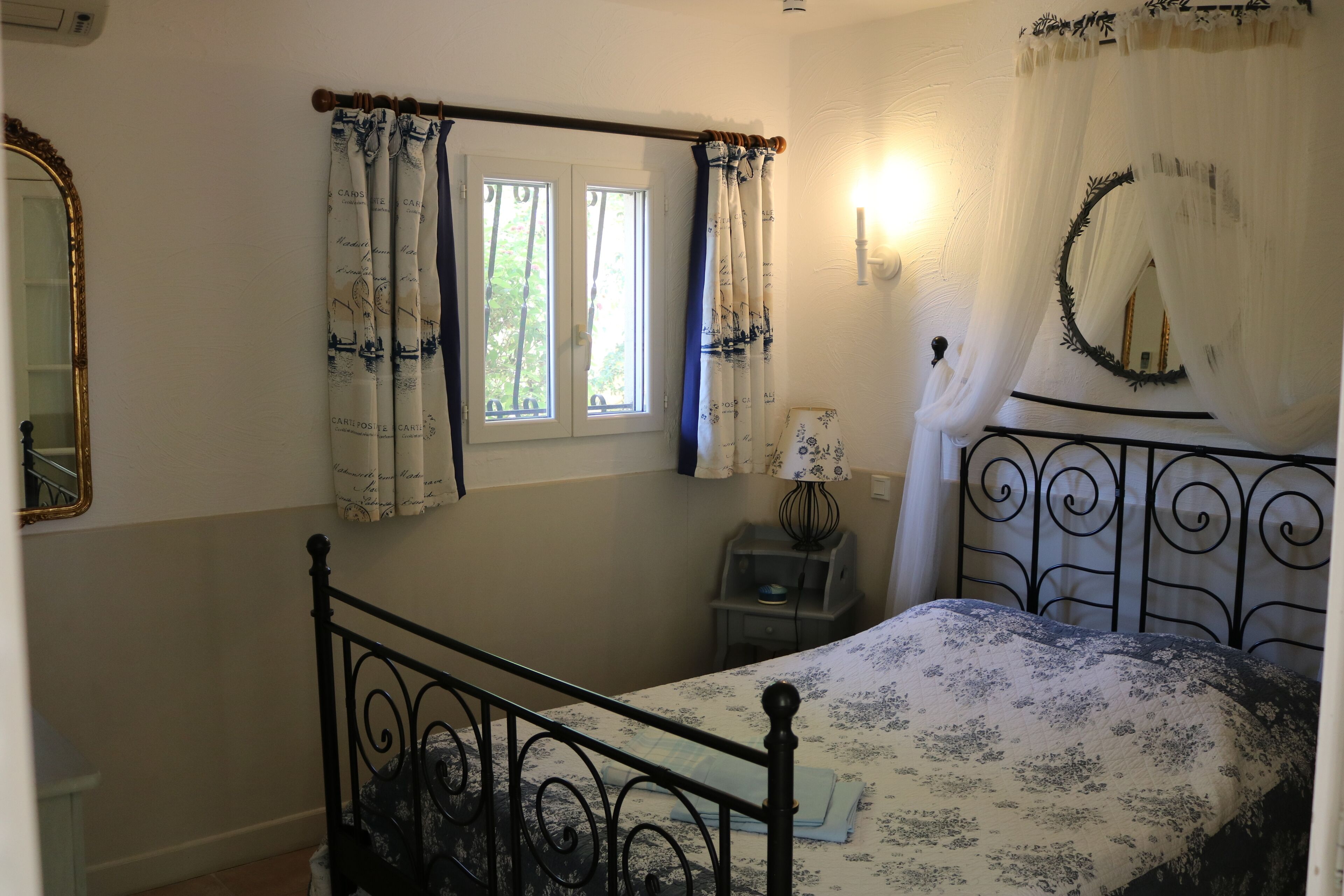 A bedroom with a bed, a window with curtains, a bedside table with a lamp, and a mirror on the wall.
