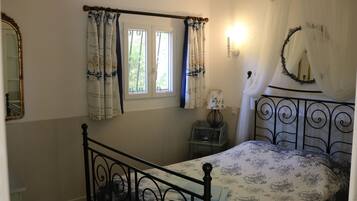 A bedroom with a bed, a window with curtains, a bedside table with a lamp, and a mirror on the wall.