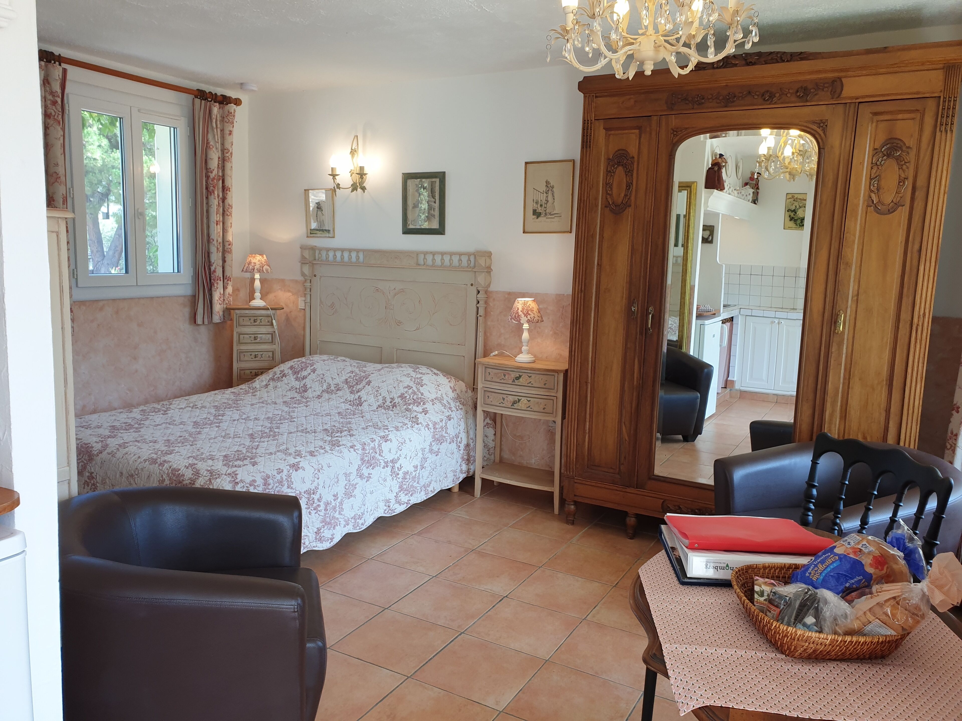A bedroom with a bed, a dresser, a chair, a mirror, and a dining area visible in the background.