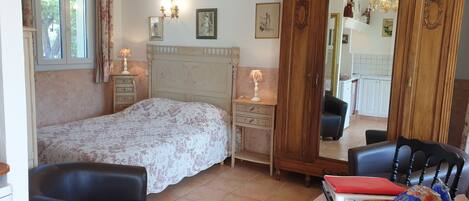 A bedroom with a bed, a dresser, a chair, a mirror, and a dining area visible in the background.