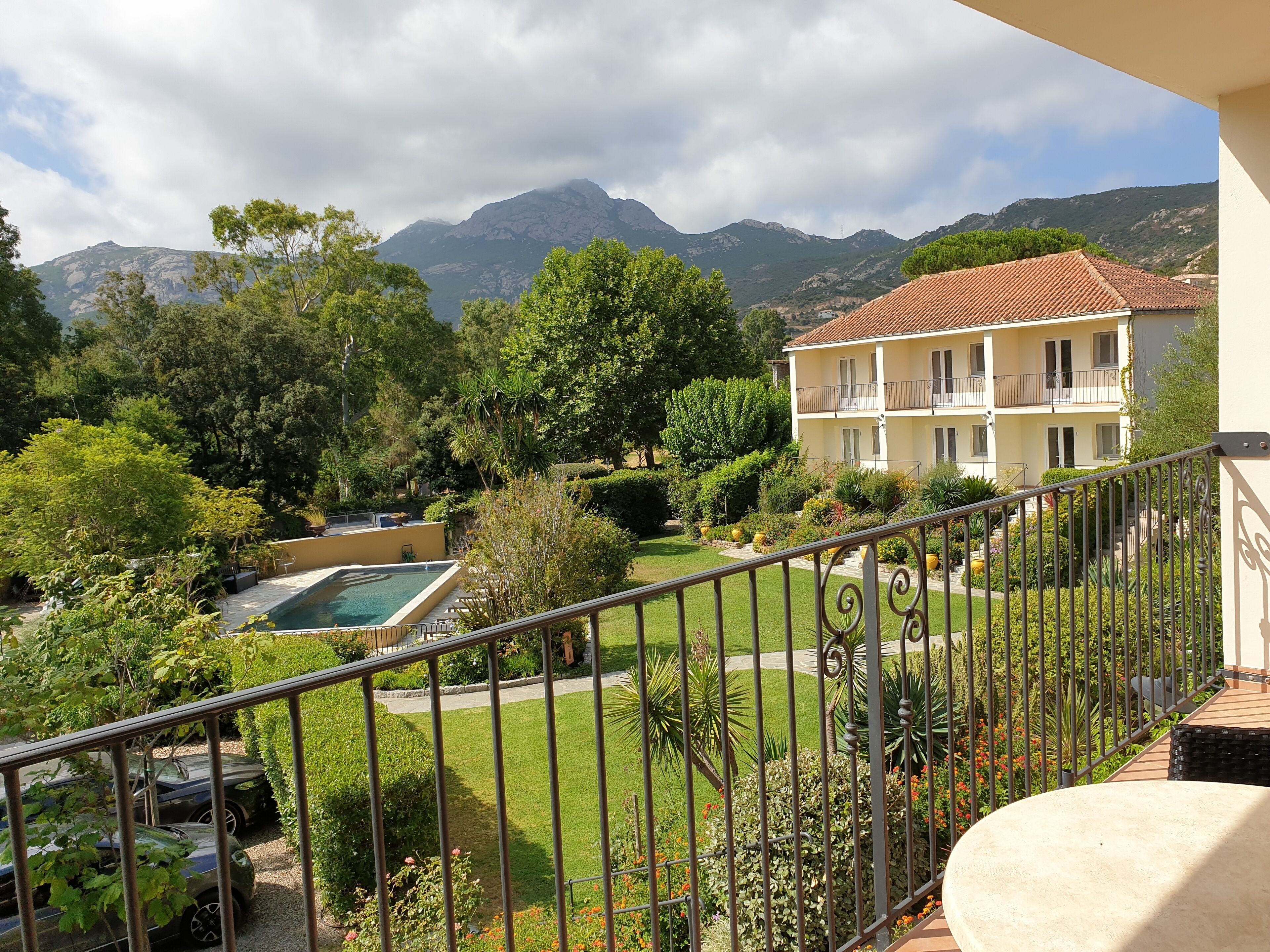 A balcony with a view of a swimming pool, lush greenery, and a building with a tiled roof.