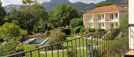 A balcony with a view of a swimming pool, lush greenery, and a building with a tiled roof.