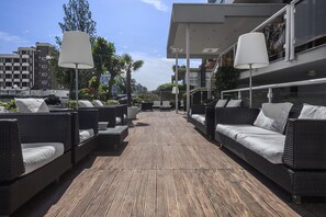Un área de lounge en la azotea con mobiliario de mimbre y cojines blancos, que cuenta con una terraza de madera y arquitectura moderna al fondo.