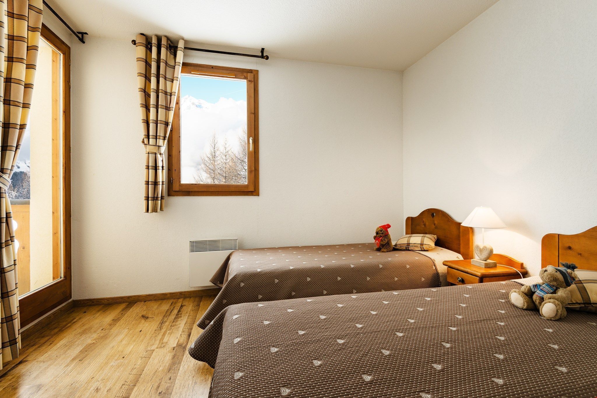 Une chambre avec deux lits, une fenêtre avec des rideaux, une table de chevet avec une lampe et un ours en peluche sur le lit de droite.